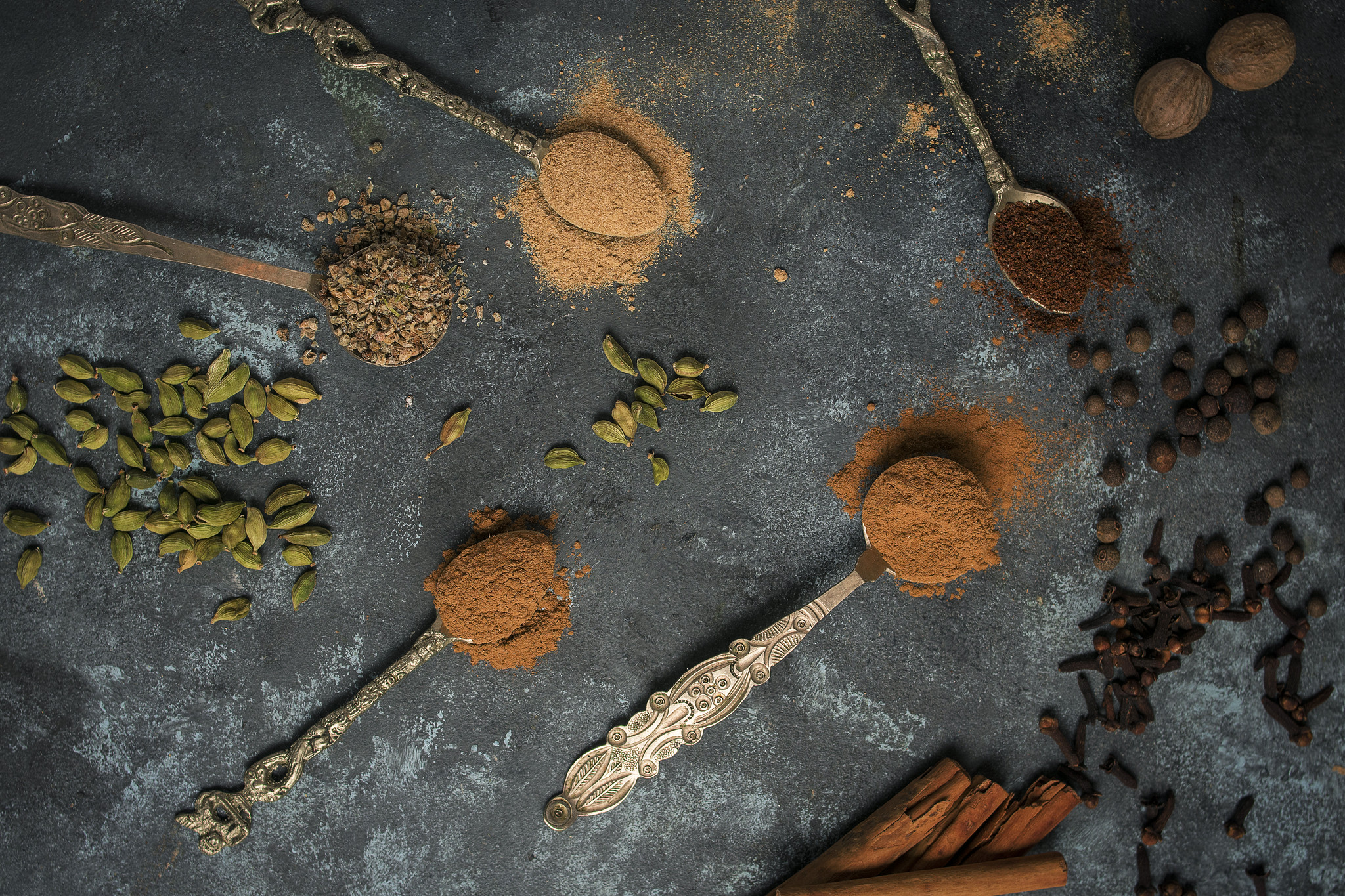 Whole and ground spices including cardamom, cinnamon, cloves and nutmeg arranged with vintage spoons on a dark surface.